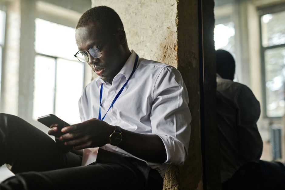 Um jovem homem afro-americano de óculos usando um smartphone dentro de casa, sentado perto de uma janela.