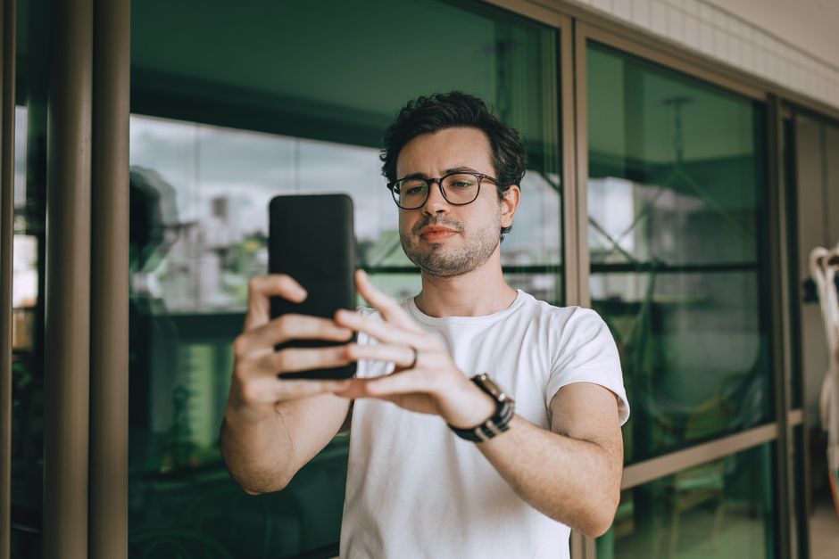 Um jovem usando óculos e uma camiseta branca tira uma selfie ao ar livre com seu smartphone.